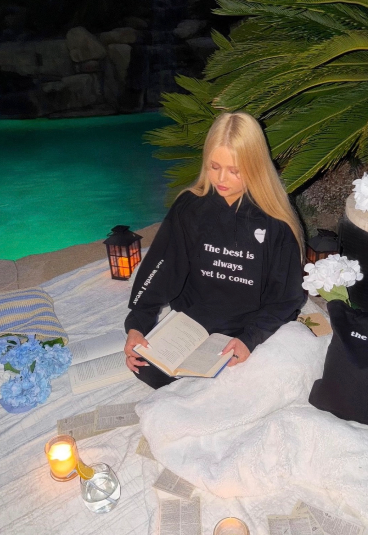 Person sitting by a pool reading a book with candles and flowers around, wearing a sweatshirt with text.