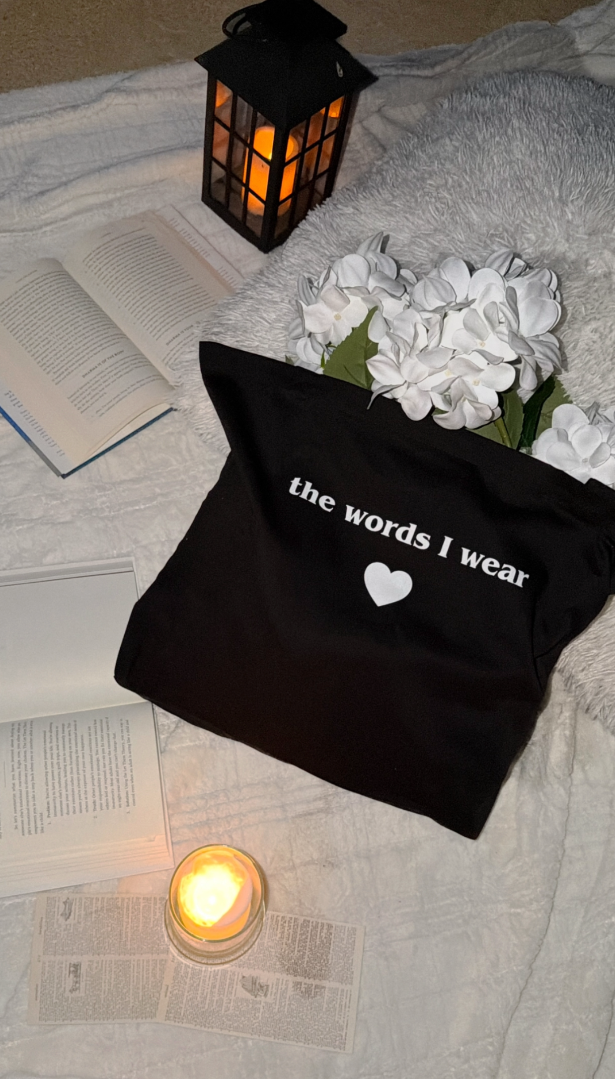 Black bag with 'the words I wear' text on a bed with flowers and a candle.